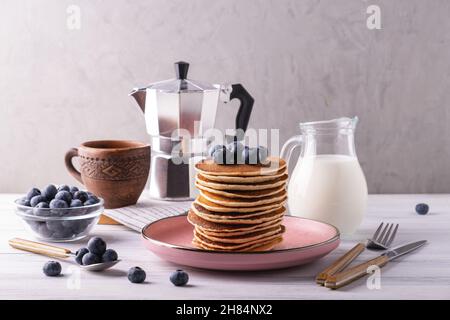 Serviert Frühstück mit Pfannkuchen auf rosafarbener Kerkerplatte, Heidelbeeren, Kaffeemaschine und Milch in Glaskanne. Selektiver und weicher Fokus. Stockfoto