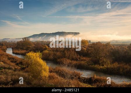 Wunderschöne Landschaft am Flussufer mit den Herbstfarben in den Bäumen und dem Nebel, der den Berg bedeckt, Torrejón de Ardoz, Madrid, Spanien Stockfoto