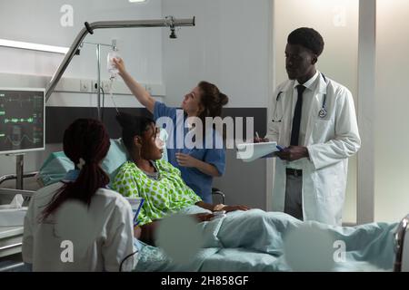 Assistentin beim Überprüfen des infusionsbeutels, während afroamerikanischer Therapeut-Arzt Krankheitssymptome analysierte und die medikamentöse Behandlung diskutierte. Kranker Patient, der sich nach der Operation im Bett erholt Stockfoto