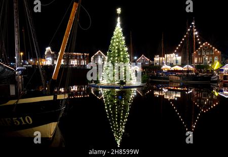 Carolinensiel, Deutschland. 27th. November 2021. Der schwimmende Weihnachtsbaum leuchtet im historischen Hafen des Dorfes. Seit 1995 wird der Baum traditionell am Samstag vor dem ersten Adventssonntag aufgesetzt und die Lichter eingeschaltet. Quelle: Hauke-Christian Dittrich/dpa/Alamy Live News Stockfoto