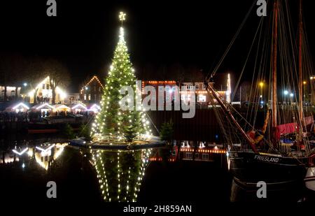 Carolinensiel, Deutschland. 27th. November 2021. Der schwimmende Weihnachtsbaum leuchtet im historischen Hafen des Dorfes. Seit 1995 wird der Baum traditionell am Samstag vor dem ersten Adventssonntag aufgesetzt und die Lichter eingeschaltet. Quelle: Hauke-Christian Dittrich/dpa/Alamy Live News Stockfoto