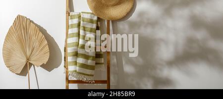 Strandtuch aus Bio-Leinen auf Holzleiter, Strohhut und getrocknetes Palmblatt an der weißen Wand mit Schatten von Baumblättern Stockfoto