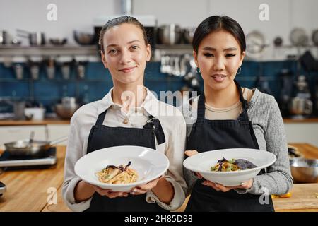 Porträt von zwei lächelnden jungen Frauen, die Schürzen tragen und Gourmet-Gerichte in der professionellen Küche halten, Kopierraum Stockfoto
