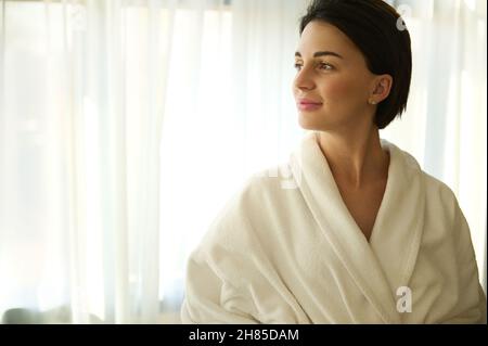 Ruhige wunderschöne schöne gepflegte junge dunkelhaarige Frau mit einem stilvollen kurzen Haarschnitt in einem weißen Frottee-Bademantel, lächelnd und Blick zur Seite aga Stockfoto