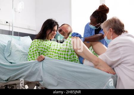 Multiethnische Geburtshilfe-Spezialisten, die ein Kind zur Verfügung stellen und Frauen bei der Wehen mit Wehen helfen. Schwanger gebärende Person mit Ehemann in der Krankenstation bei der Entbindungszeit Stockfoto