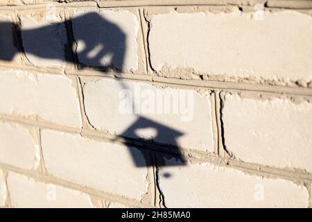 Unschärfe Schatten der Hand mit Weihnachtsspielzeug kleines Haus auf weißen Ziegelwand. Licht- und Schattenspielzeug. Hausschatten mit Spiegelung an der Hauswand, outd Stockfoto