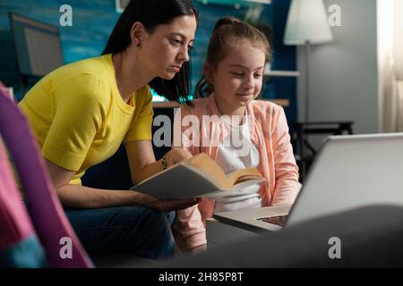 Mutter hält ein Literaturbuch in den Händen und hilft ihrer Tochter bei den Schulaufgaben, die sie im Wohnzimmer auf einer großen Bohne sitzt. Schulkind, das für eine akademische Prüfung mit einer elearning-Lernplattform studiert Stockfoto
