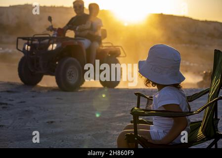 Eine selektive Fokusaufnahme eines Kindes, das auf einem Stuhl sitzt. Zwei Personen fahren mit dem Quad in der Wüste. Stockfoto