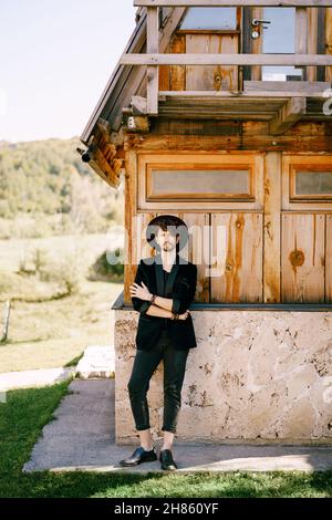 Ein junger Mann mit Hut steht in der Nähe eines alten Holzhauses Stockfoto