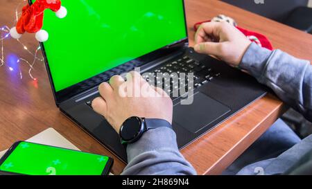 Nahaufnahme Mann Hände tippen auf Laptop-Tastatur mit leerem grünen Bildschirm, Smartphone-Mockup und Weihnachtsdekoration auf dem Hintergrund. Urlaubskonzept. Büro Stockfoto