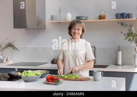 Glückliche ältere Dame, die am Tisch frisches Gemüse für Salat hackt Stockfoto