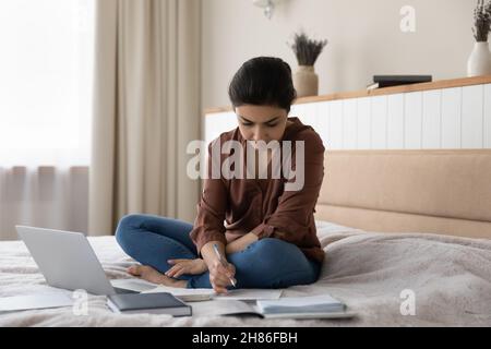 Konzentrierte junge indische Frau, die entfernt im Schlafzimmer studiert. Stockfoto