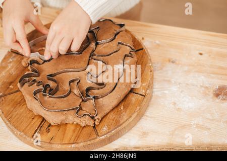 Kinderhände schnitzen Kekse auf Lebkuchenteig aus Figuren. Mädchen bereitet weihnachtskekse auf einem Holztisch zu Stockfoto