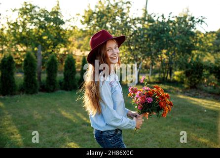Ein rothaariges Teenager-Mädchen in einem roten Hut tanzt auf dem Rasen mit einem Blumenstrauß Stockfoto