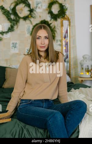 Porträt der jungen schönen kaukasischen Frau auf dem Bett im Dekorationsraum für Weihnachtsferien sitzen. Stockfoto