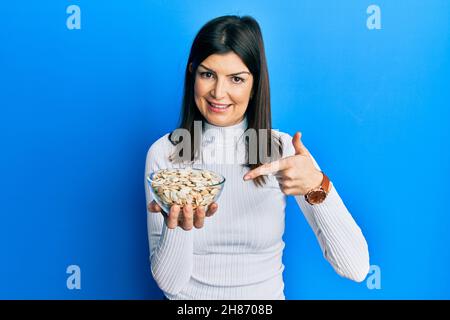 Junge hispanische Frau hält Kürbiskerne Schüssel lächelnd glücklich zeigt mit Hand und Finger Stockfoto