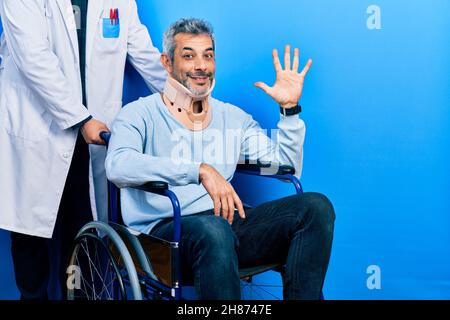 Ein hübscher Mann mittleren Alters mit grauen Haaren auf dem Rollstuhl, der einen Halsband trägt und mit den fünf Fingern nach oben zeigt, während er selbstbewusst lächelt Stockfoto