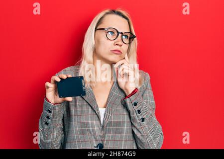 Schöne kaukasische blonde Geschäftsfrau hält ssd-Speicher ernstes Gesicht denken über Frage mit Hand auf Kinn, nachdenklich über verwirrende Idee Stockfoto