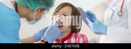 Otorhinolaryngologe Arzt in schützende medizinische Maske untersucht Hals des kleinen Mädchens mit Spatel in der Klinik Stockfoto