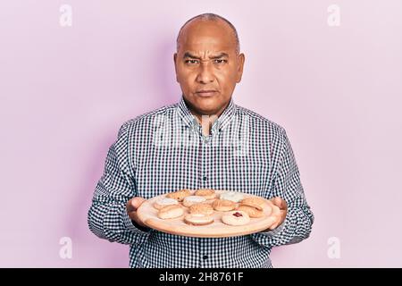 lateinischer Mann mittleren Alters hält Tablett mit Keksen skeptisch und nervös, runzelte die Stirn wegen des Problems. Negative Person. Stockfoto