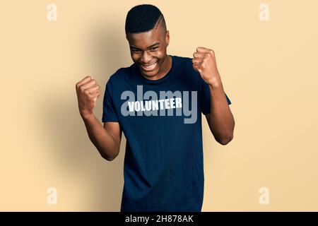 Der junge afroamerikanische Mann im T-Shirt des Freiwilligen war sehr glücklich und aufgeregt, als er mit erhobenen Armen die Siegergeste machte, lächelte und nach Erfolg schrie. Stockfoto