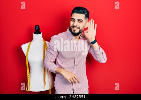 Hübscher Schneider mit Bart, der neben dem menschlichen Darstellungsobjekt steht, lächelt mit der Hand über dem Ohr und hört Gerüchte oder Klatsch. Taubheitskonzept. Stockfoto