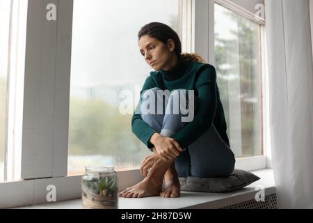 Traurige hispanische Dame sitzt barfuß auf dem Fensterbrett fühlen sich abwesend gesinnt Stockfoto