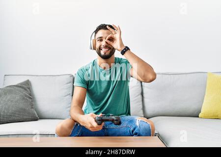 Gutaussehender hispanischer Mann mit Kopfhörern, der Videospiel hält, der Controller lächelt und ein Schild mit der Hand auf dem Auge durch die Finger schaut Stockfoto