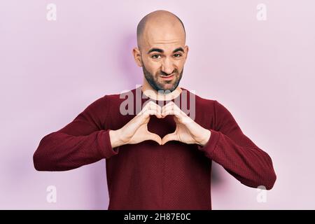 Junger kahler Mann tut Herz-Symbol mit Händen ahnungslos und verwirrt Ausdruck. Zweifel Konzept. Stockfoto