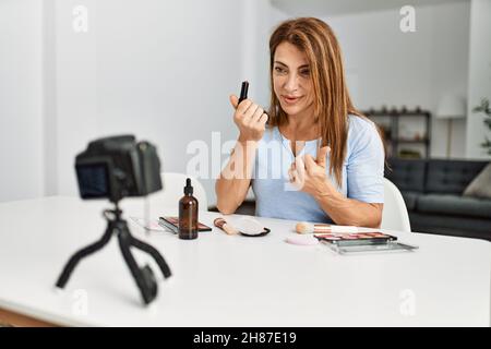Kaukasische Frau mittleren Alters macht Make-up-Tutorial mit Kamera sitzen auf dem Tisch zu Hause. Stockfoto