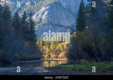 Yosemite-Nationalpark, Kalifornien, USA. Blick auf den ruhigen Merced River in der Nähe von Yosemite Village, herbstliche, hohe Granitklippen, die sich über dem Hotel erheben. Stockfoto