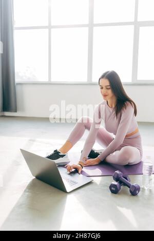 Junge sportliche Frau trägt Sportkleidung, die online Fitness-Yoga-Kurs mit einem Laptop-Computer sucht, der zu Hause auf einer Matte sitzt. Stockfoto