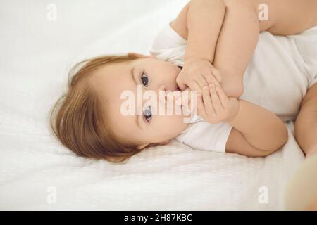 Schönes, unbeschwertes Mädchen von sechs Monaten liegt auf weißer Bettwäsche und spielt mit ihren Beinen. Stockfoto