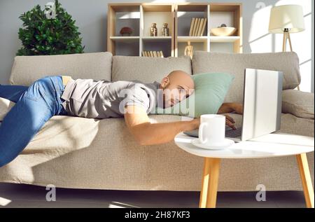 Gelangweilter, depressiver, fauler junger Mann, der auf der Couch lag und seinen Laptop ansah Stockfoto