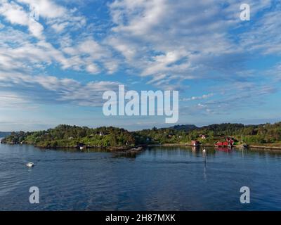 Ein kleines Motorboot, das an einem hellen Summers-Morgen im Juni an der bewaldeten Küste mit verstreuten Häusern und Hütten im Bergen Fjord vorbeifährt. Stockfoto