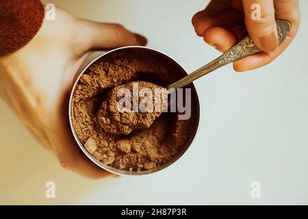 Eine Frau in einer braunen Jacke hält ein Glas duftenden Kakaopulvers in ihren Händen und nimmt mit einem Teelöffel eine Portion Kakao daraus heraus. Draufsicht. Die PR Stockfoto