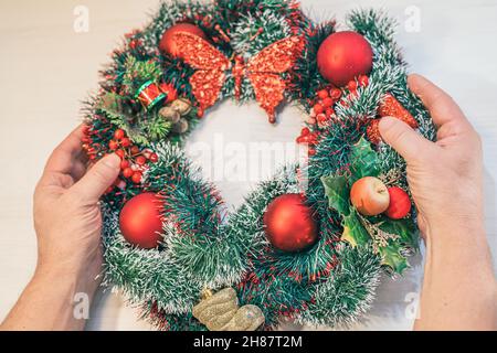 Weihnachtskranz aus Lametta und rote Weihnachtsdekoration an der Haustür. Draufsicht auf die Dekoration der männlichen, gealterten Hände Stockfoto