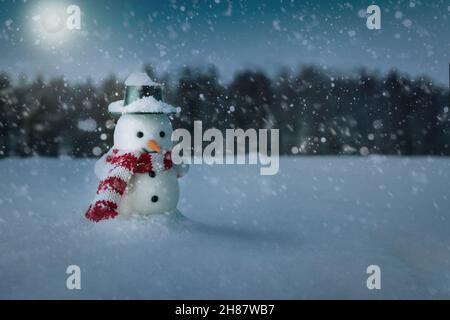 Schneemann mit Hut und Schal, der nachts auf einem schneebedeckten Feld in der Nähe des Waldes steht Stockfoto