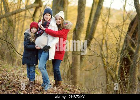 Eine junge, schwanger gebliebene Frau umarmte ihre älteren Töchter. Ältere Geschwister haben Spaß mit ihrer Schwangeren Mutter im Freien. Mutter und ihre Kinder verbringen viel Zeit Stockfoto