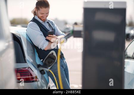 Junger lächelnder Mann mit Smartphone in der Nähe eines Elektroautos an der Ladestation Stockfoto