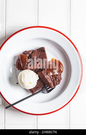 Vegane Brownie-Quadrate mit einer Kugel milchfreies Eis und Apfelkaramell auf einem weißen Teller. Pflanzliches Diät-Konzept. Stockfoto