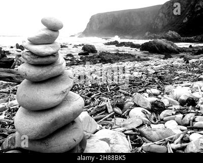 Ein abgeschiedener Strand mit Kieselsteinen und Treibholz, mit einem Turm aus Kieselsteinen übereinander gestapelt im Vordergrund, in schwarz und weiß, entlang der Th Stockfoto