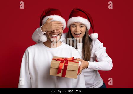 Weihnachtsüberraschung. Glückliche junge Frau hält eine Geschenkbox und deckt die Augen des Freundes ab Stockfoto