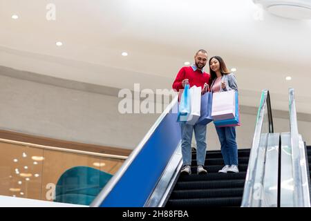 Die ganze Länge eines jungen interracial Paares, das auf einer beweglichen Treppe in der Mall hinuntergeht, in Einkaufstaschen schaut, in leeren Raum Stockfoto