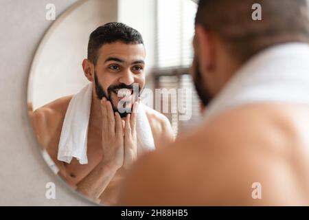 Selbstbewusster junger Araber, der im Spiegel schaut und den Bart berührt Stockfoto