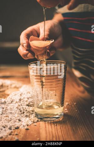 Frau trennt Eigelb von Eiweiß in Glas. Teig auf Holztisch machen. Zubereitung von Speisen in der Küche. Getöntes Bild Stockfoto