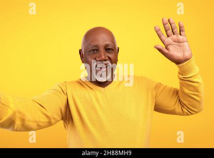 Glücklicher älterer schwarzer Mann, der Selfie macht und vor der Kamera auf orangefarbenem Studiohintergrund winkt Stockfoto