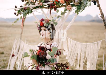Die Braut in einem Kranz mit einem Blumenstrauß steht neben dem Hochzeitsbogen auf dem Feld Stockfoto