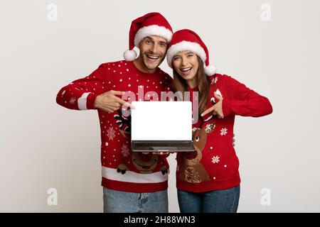 Fröhliches Junges Paar, Das Weihnachtshützen Trägt Und Auf Laptop Mit Leerem Bildschirm Zeigt Stockfoto