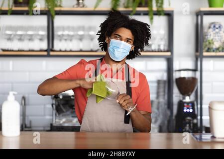 Arbeit Während Der Pandemie. Junger schwarzer Barkeeper mit medizinischer Maske, der an der Theke steht Stockfoto